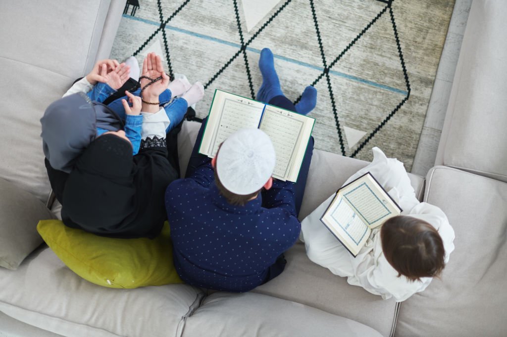 Top view of young muslim family reading Quran during Ramadan. Parents and kids worshiping to God, in islamic clothes at modern home