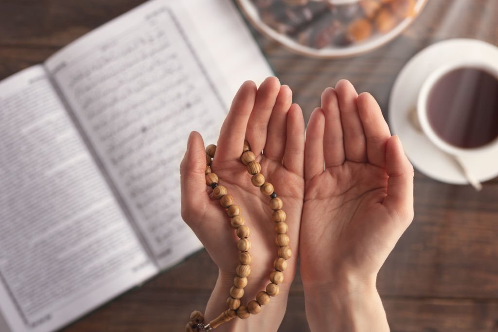 female hand of prayer with wooden beads in sunlight, iftar concept, Ramadan month, Koran, plate of dried fruit, Cup of tea on wooden table